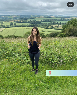 A white female with long brown hair smiles at the camera on a grassy hiking path. 